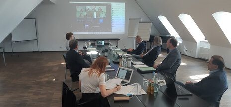 Foto der am Treffen beteiligten Projektpartner vor der Videowand mit den zugeschalteten Projektpartnern